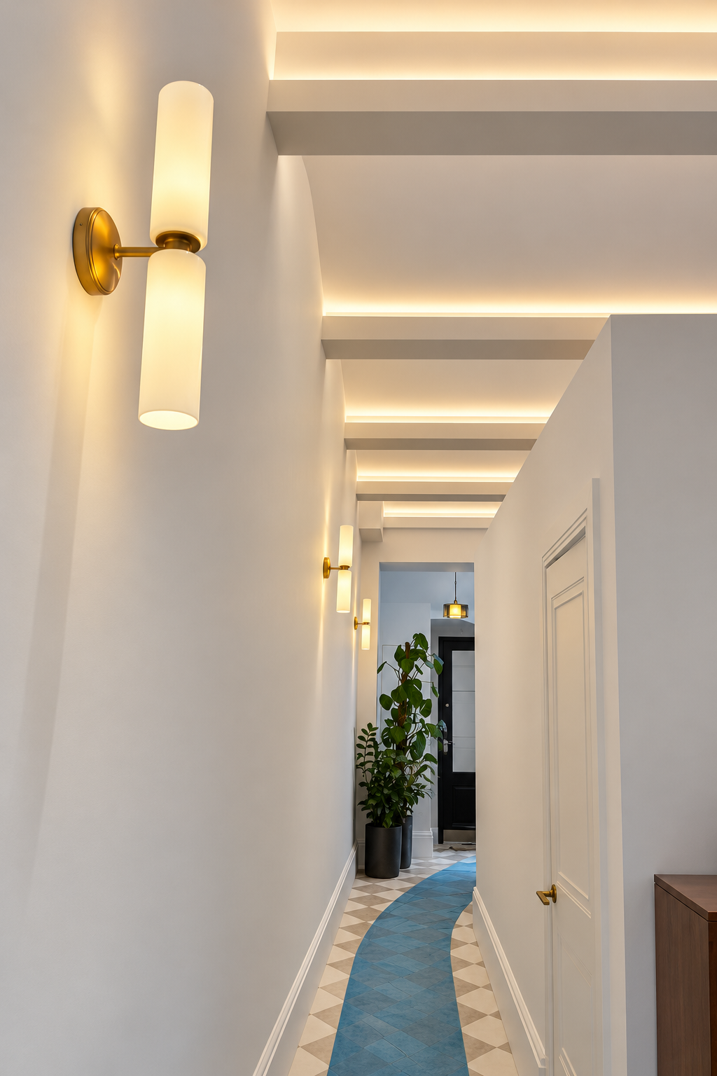White hallway with glowing brass cylinder wall lights and recessed cove lighting along coffered ceiling beams.