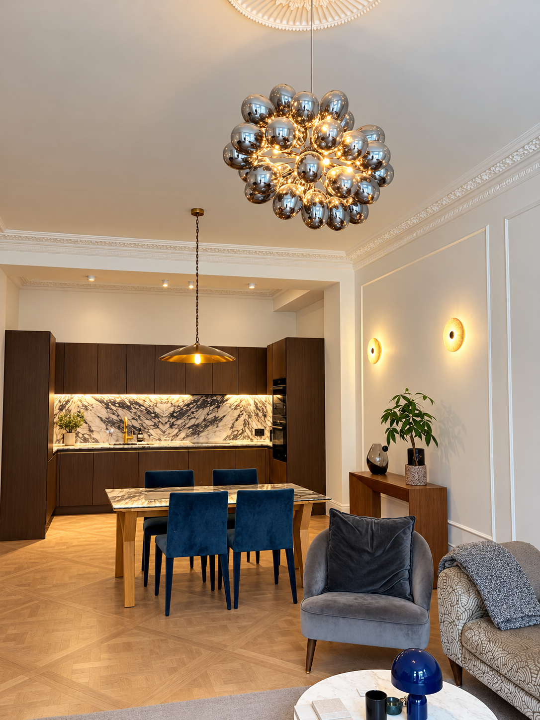Open-plan kitchen, dining and living room with a chrome bubble chandelier, blue velvet dining chairs, a dark wood kitchen with marble splashback and glowing wall lights.