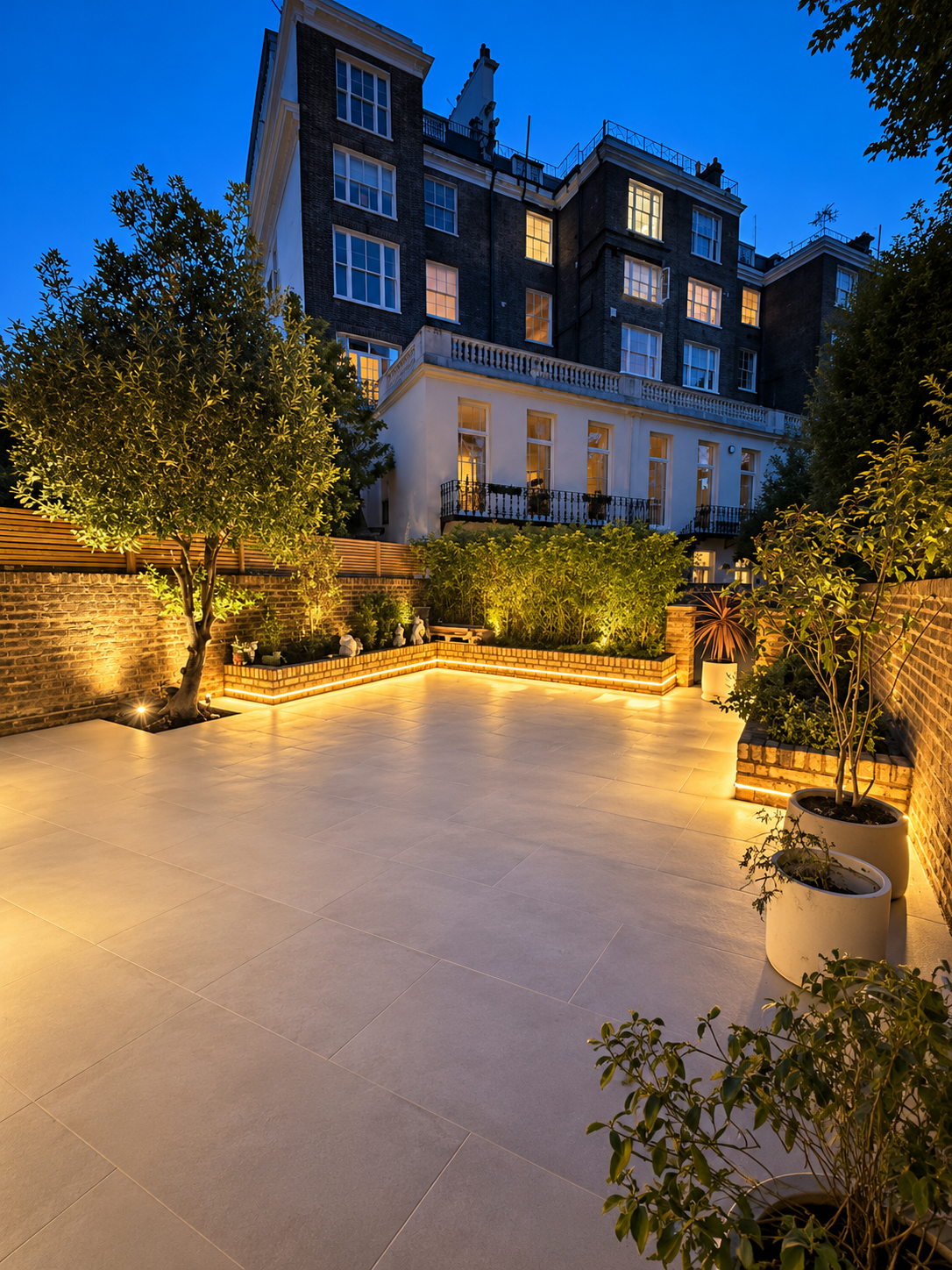 Paved garden patio at twilight with warm LED strip lighting beneath brick planters and uplights in front of a townhouse.